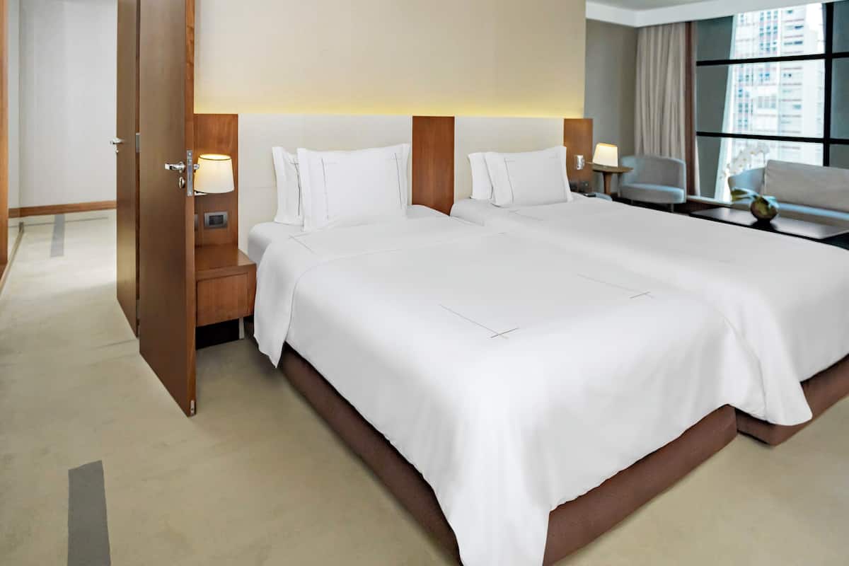 A hotel room with two beds, a nightstand with a lamp, a chair, and a view of the city through large windows.