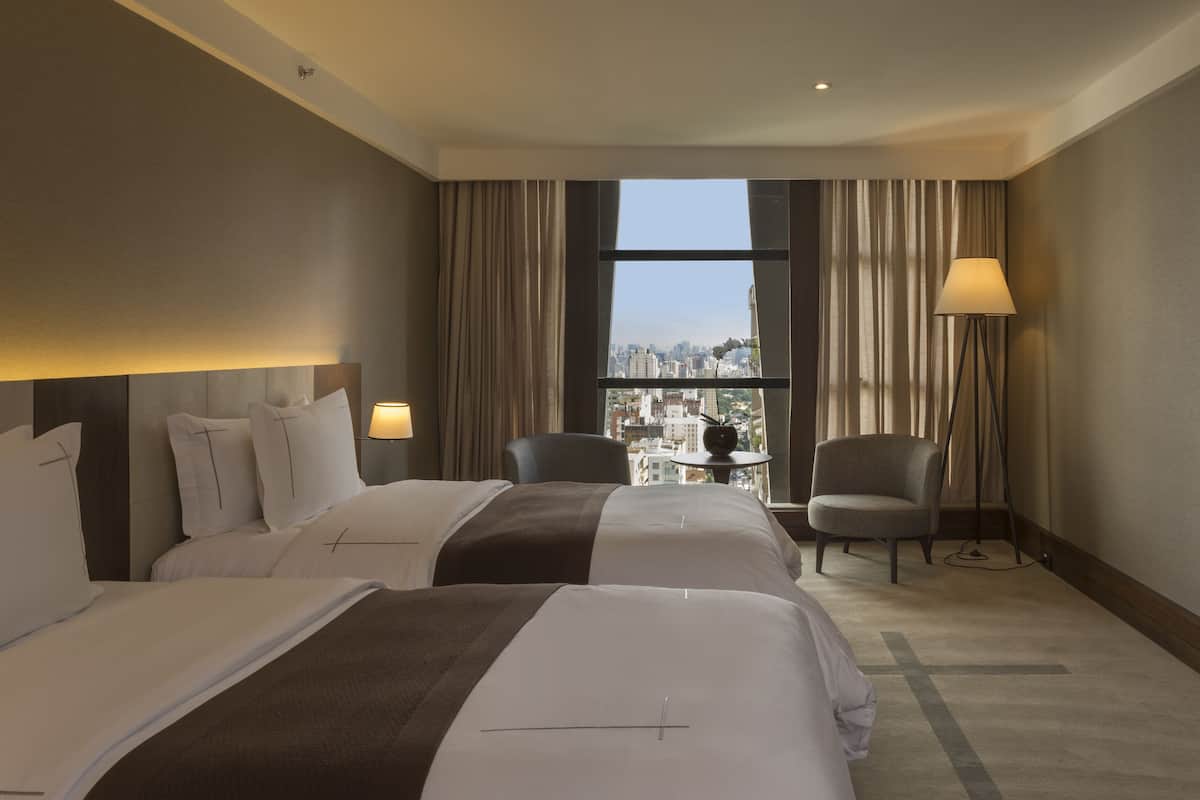 A hotel room with two beds, a chair, a TV, and a view of the city.