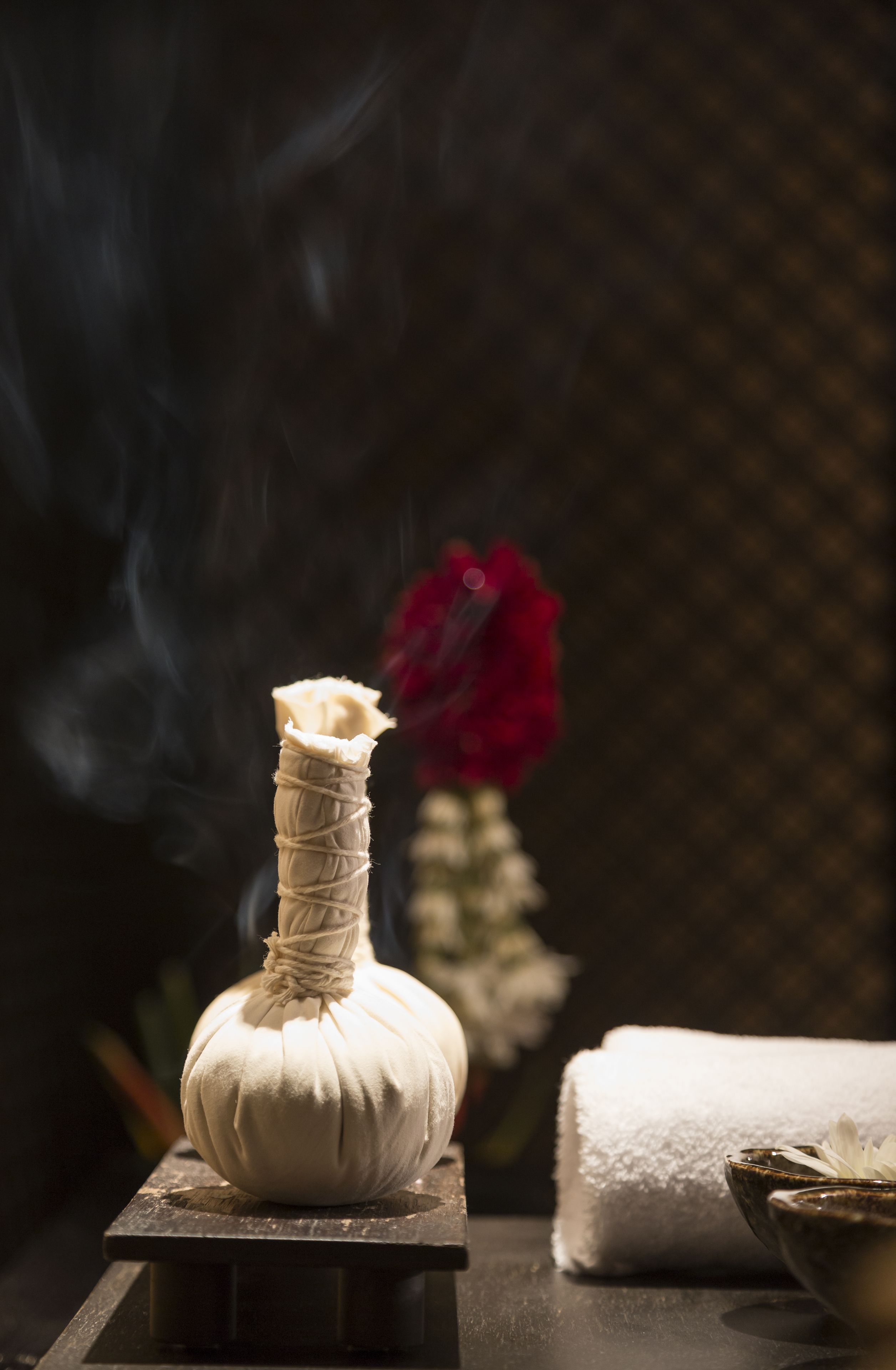 Um difusor com fumaça, uma toalha branca e uma flor vermelha ao fundo.