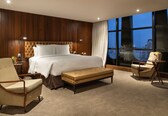 A hotel room with a large bed, a sitting area with a bench, a chair, and a small table with a vase.