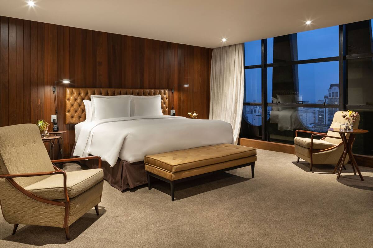 A modern hotel room with a large bed, a sitting area with a chair and a small table, a view of the city through large windows, and a wooden wall paneling.