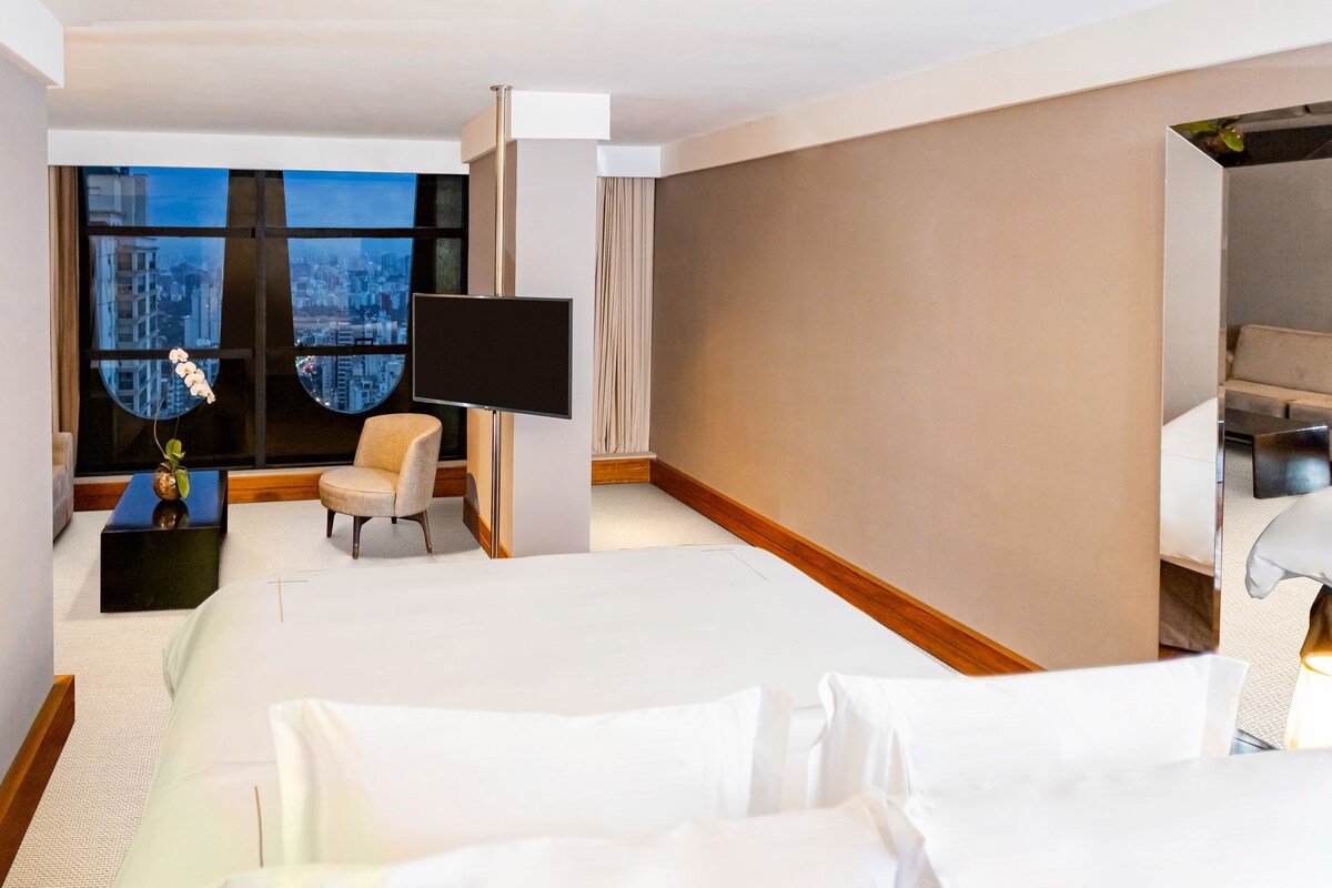 Habitación de hotel con una cama grande, televisión, una silla y vistas a la ciudad a través de amplios ventanales.