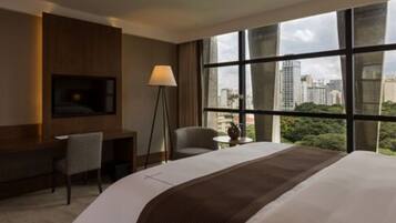 A hotel room with a large bed, a desk, a chair, a TV mounted on the wall, and a window with a city view.