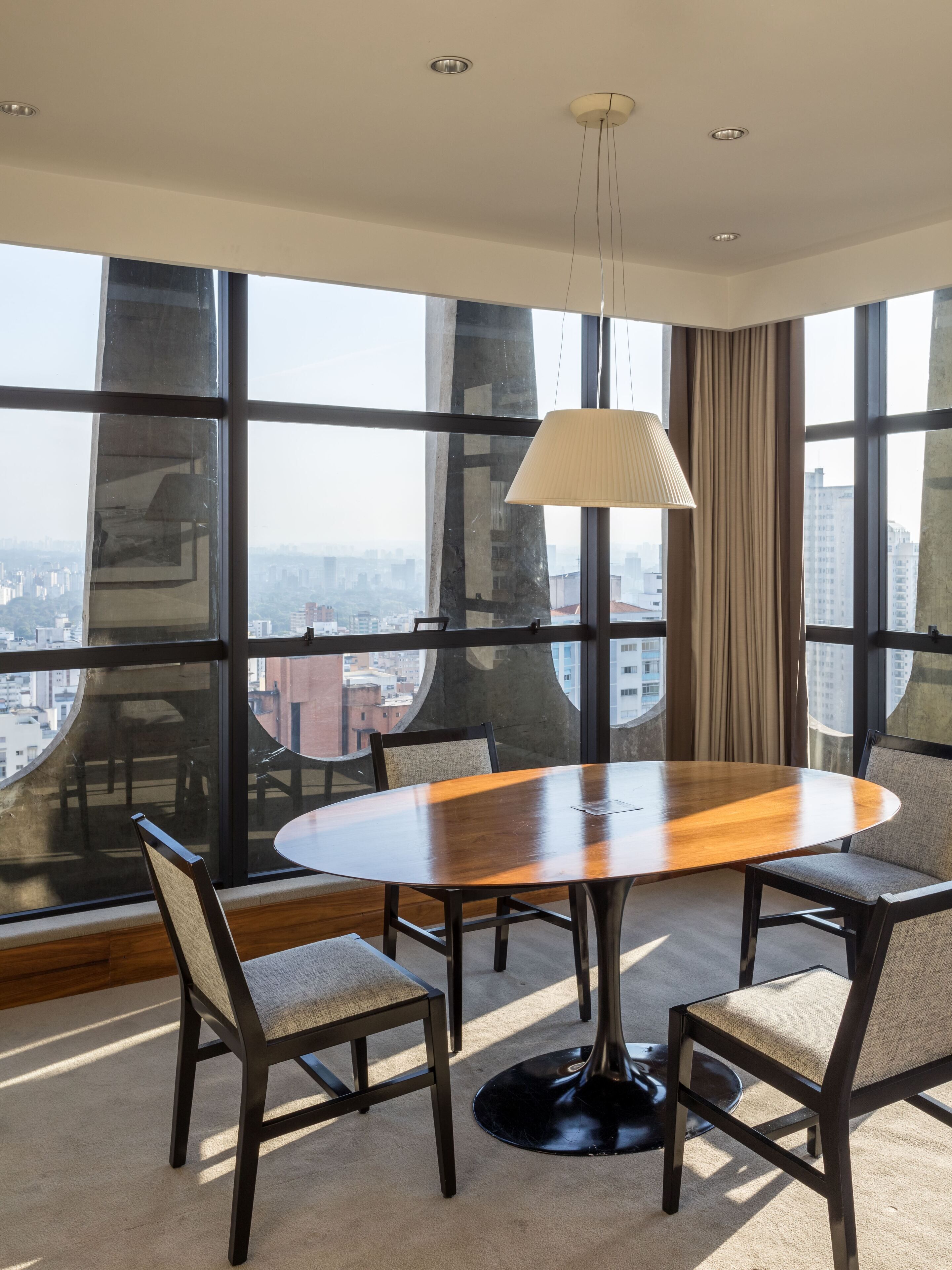 Uma sala de conferências moderna com uma mesa redonda, quatro cadeiras, uma luminária pendente e grandes janelas com vista para a cidade.
