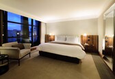 A hotel room with a large bed, a sofa, a small table, and a large window offering a city view.