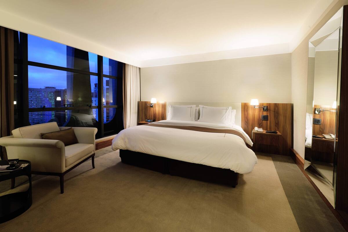 A hotel room with a large bed, a sofa, a small table, and a large window offering a city view.