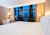 A hotel room with a large bed, a chair, two lamps, and a view of a cityscape through large windows.
