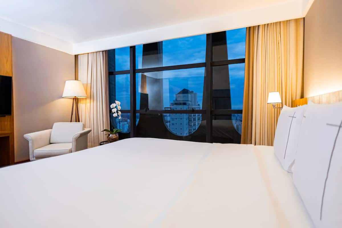 A hotel room with a large bed, a chair, two lamps, and a view of a cityscape through large windows.