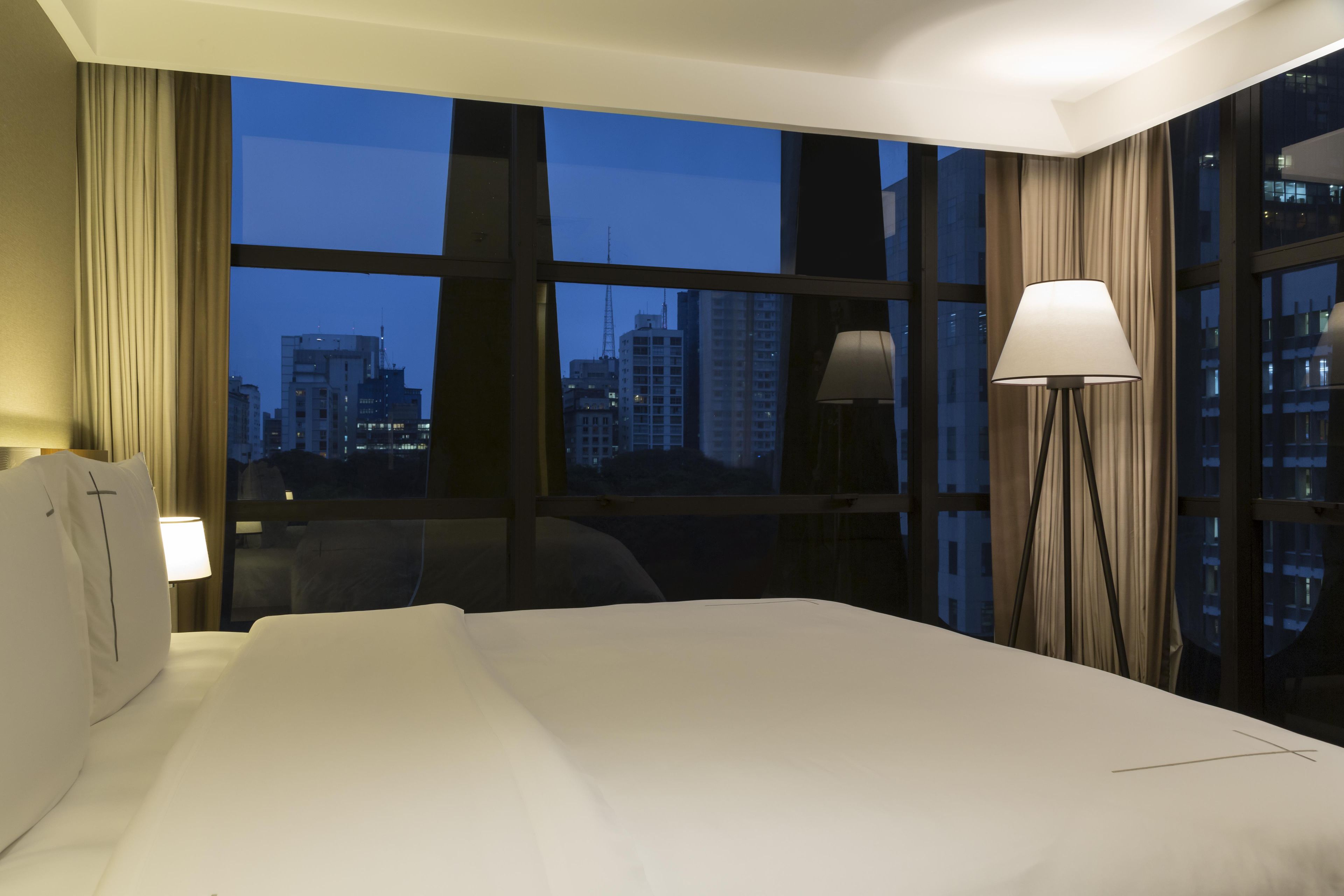 Quarto de hotel com uma cama grande, escrivaninha, cadeira, abajur e vista panorâmica da paisagem urbana através de amplas janelas.
