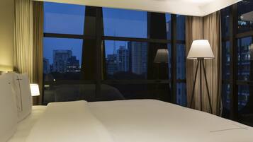 Quarto de hotel com uma cama grande, escrivaninha, cadeira, abajur e vista panorâmica da paisagem urbana através de amplas janelas.