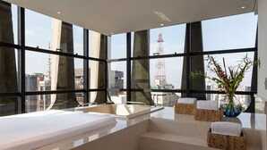 Una habitación de hotel moderna con un ventanal grande, una bañera y una pequeña zona de estar con cojines.