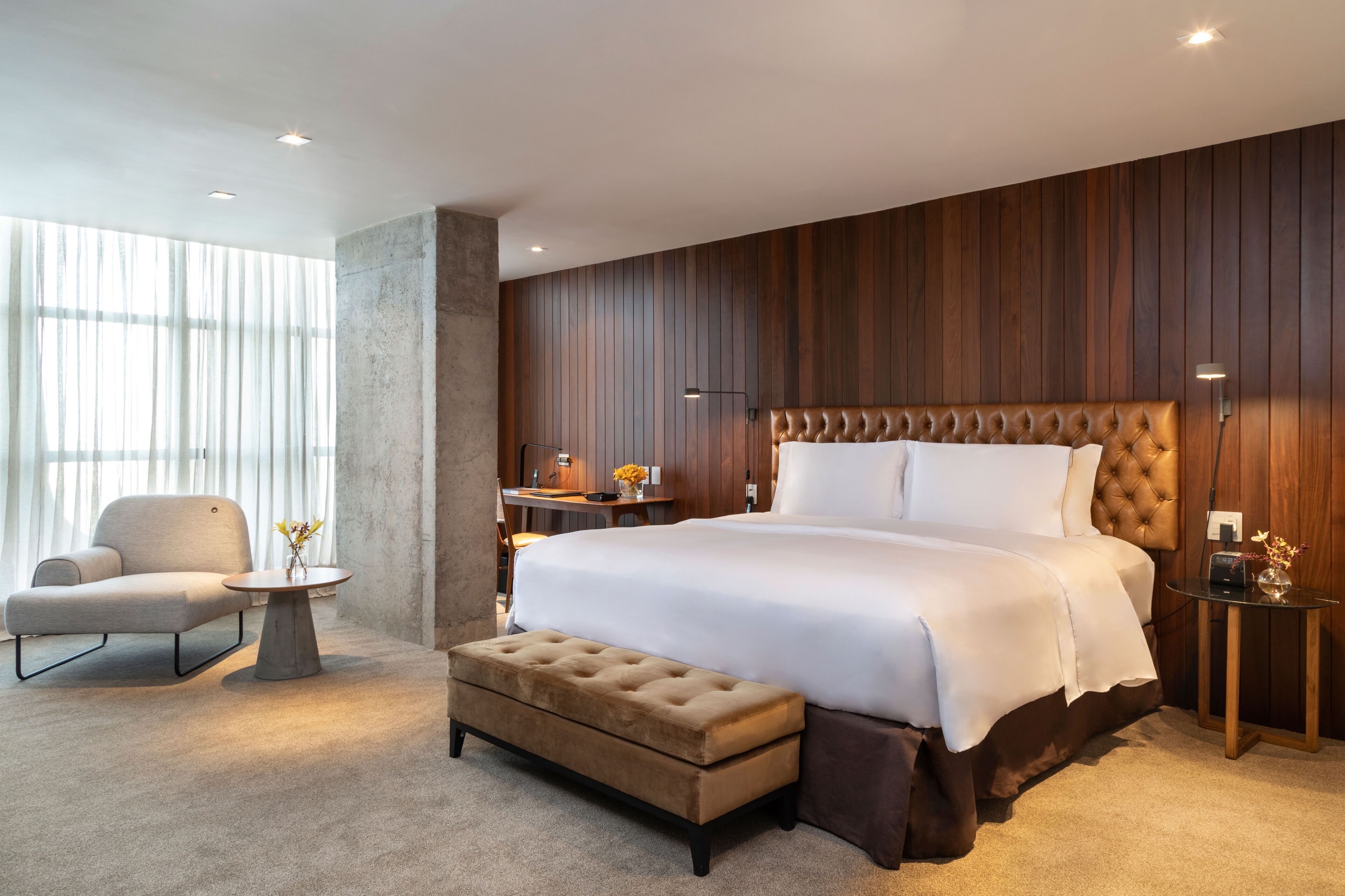 A modern bedroom with a large bed, a sitting area with a chair and table, and a wooden panel wall.