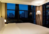 A hotel room with a large bed, a desk, a chair, a lamp, and a view of a cityscape through large windows.