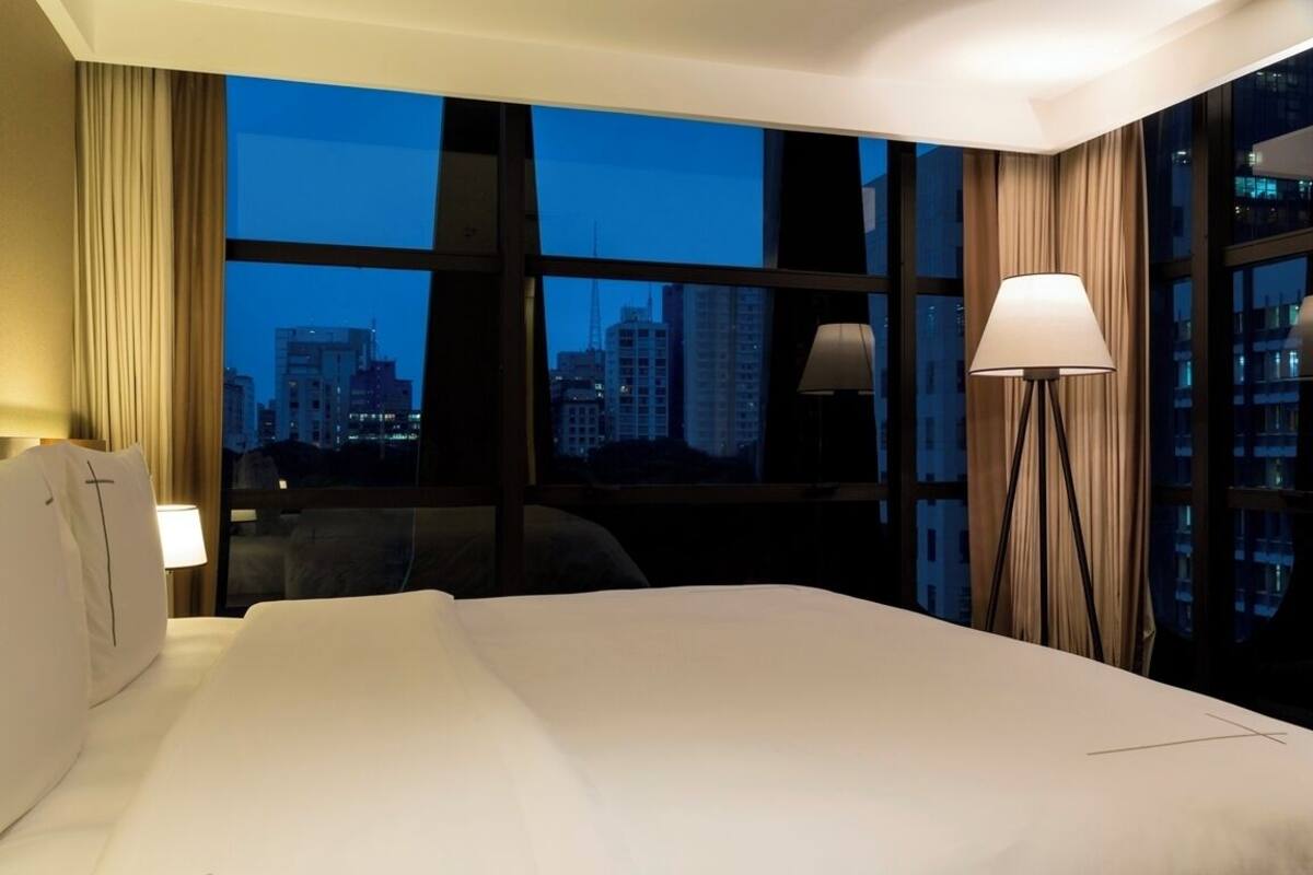 A hotel room with a large bed, a desk, a chair, a lamp, and a view of a cityscape through large windows.