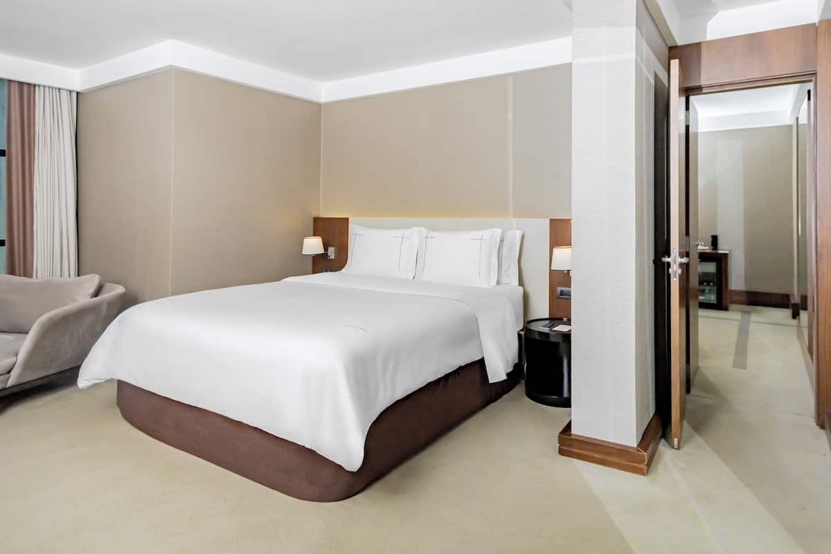 A hotel room with a large bed, a bedside table with a lamp, a chair, and a door leading to another room.