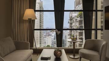 Ein Hotelzimmer mit Sofa, Ohrensessel, Couchtisch, Lampe und Blick auf hohe Gebäude.