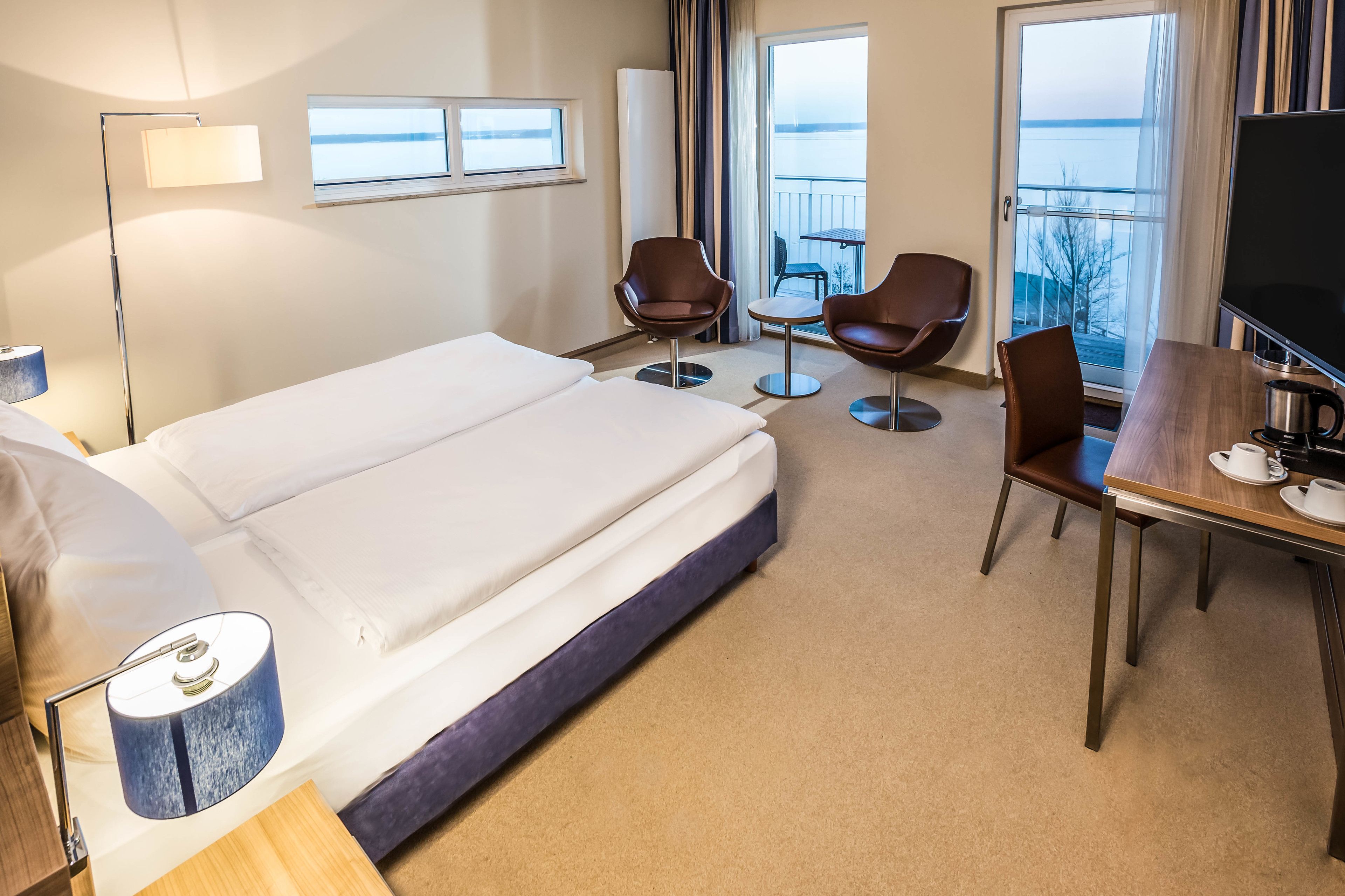 Ein Hotelzimmer mit einem großen Bett, einem Schreibtisch mit Fernseher, zwei Stühlen und Blick auf das Meer durch die Fenster.