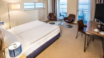 Ein Hotelzimmer mit einem großen Bett, einem Schreibtisch mit Fernseher, zwei Stühlen und Blick auf das Meer durch die Fenster.