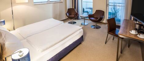 A hotel room with a large bed, a desk with a television, two chairs, and a view of the sea through the windows.