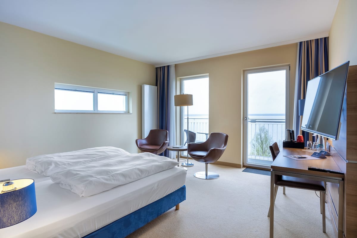 A hotel room with a bed, two chairs, a desk, a TV, and large windows.