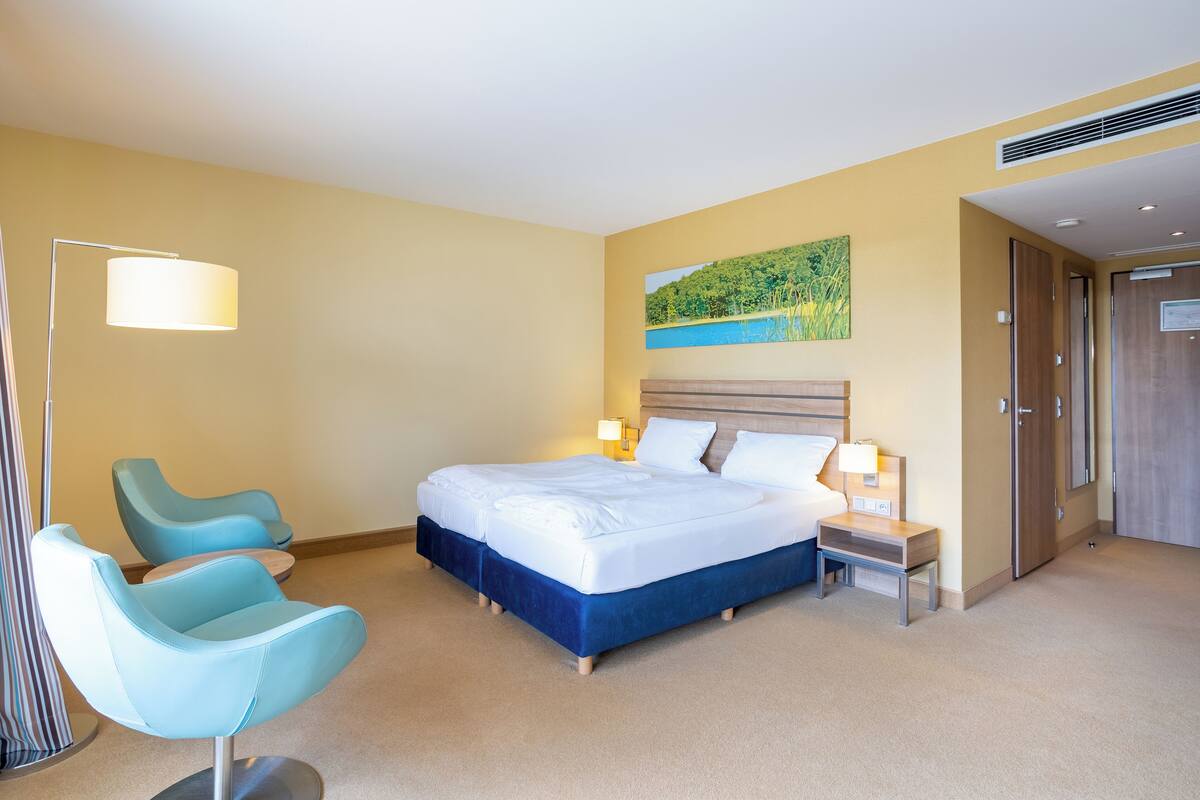A hotel room with a large bed, two bedside tables, a nightstand, a chair, a floor lamp, and a painting on the wall.