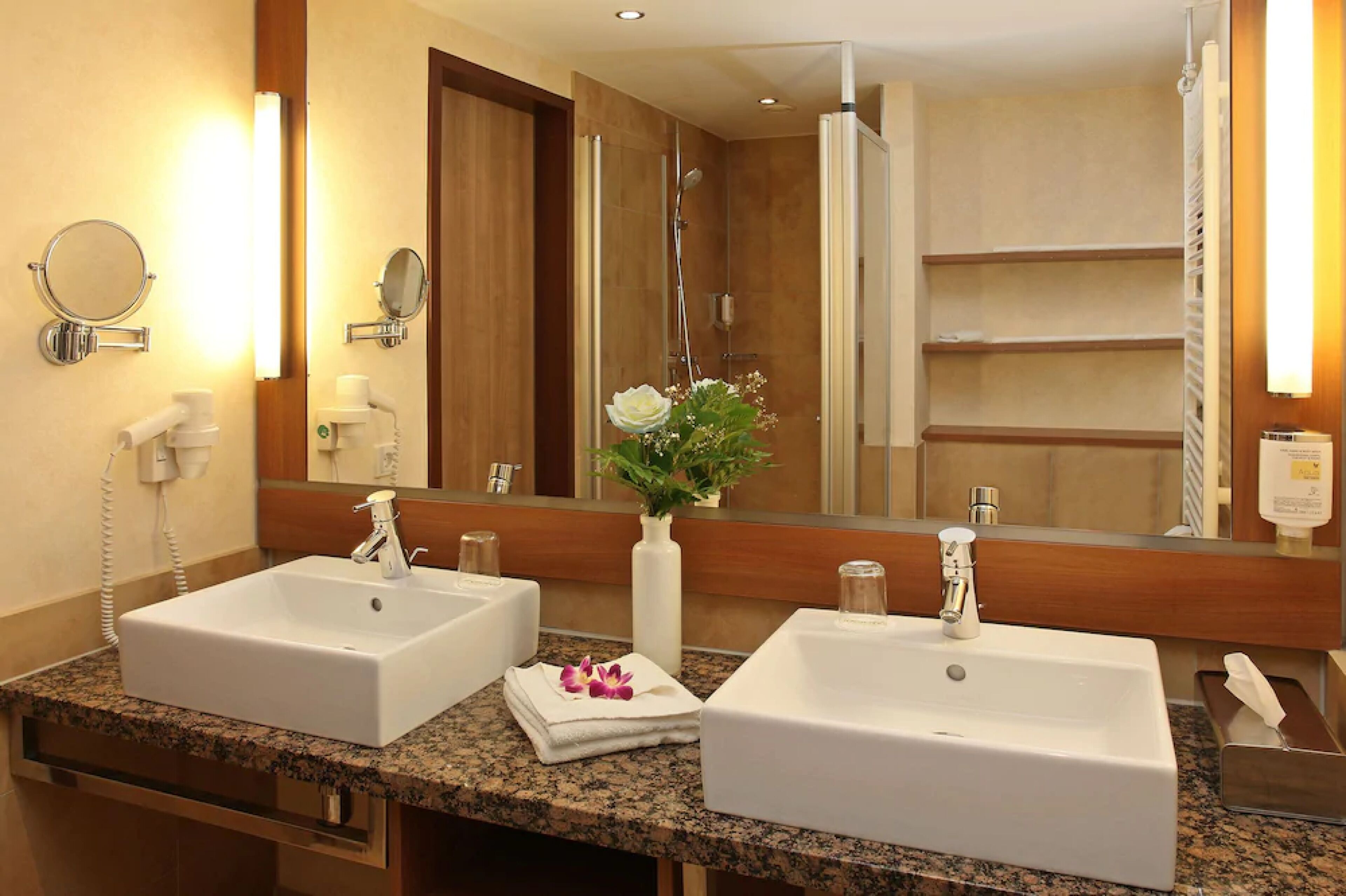 Un baño con dos lavabos, un espejo, ducha y un florero con flores.