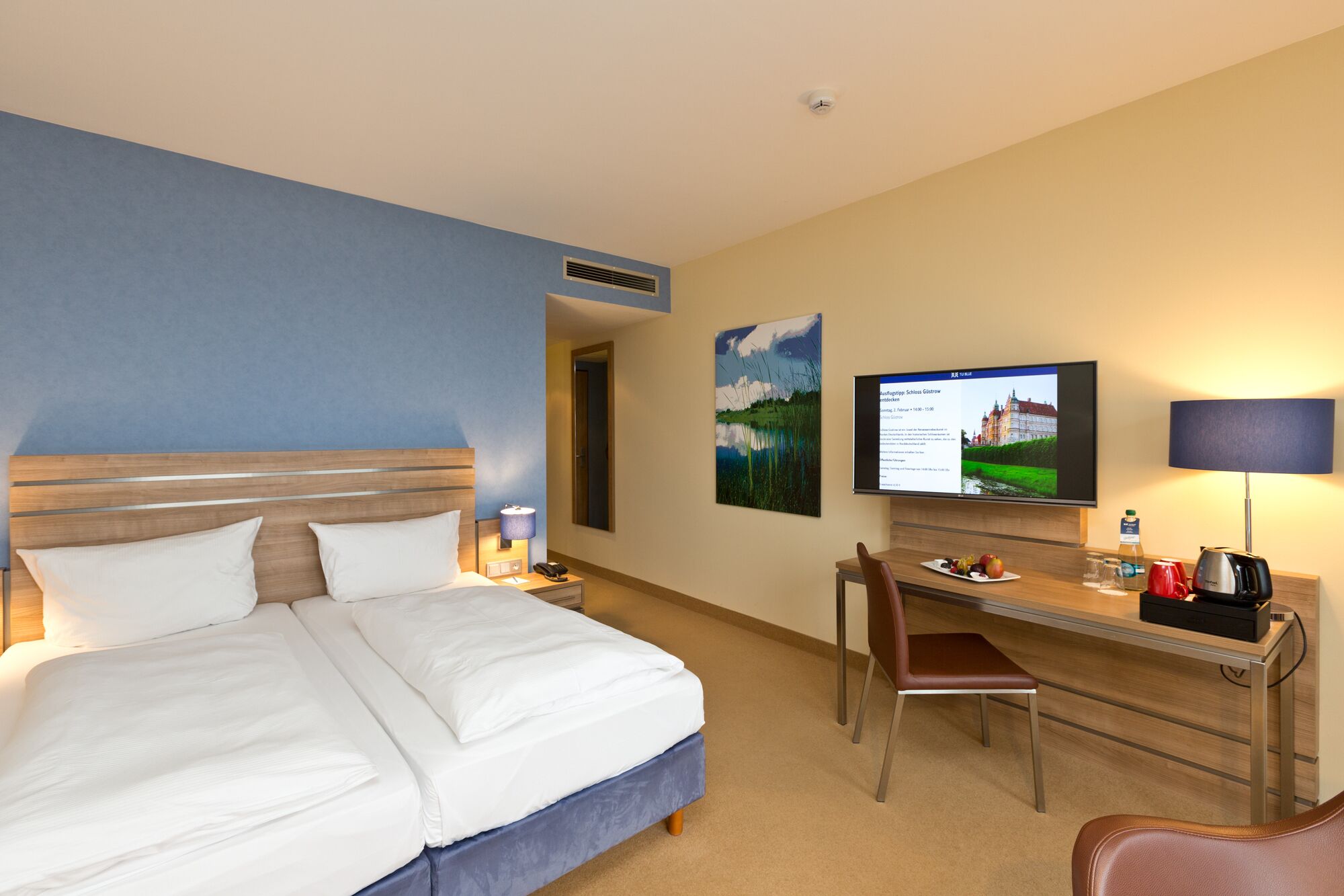Habitación de hotel con cama, escritorio, silla, televisor y una obra de arte colgada en la pared.