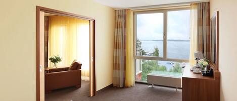 A hotel room with a bed, a sofa, a desk with a lamp, and a large window offering a view of the sea.