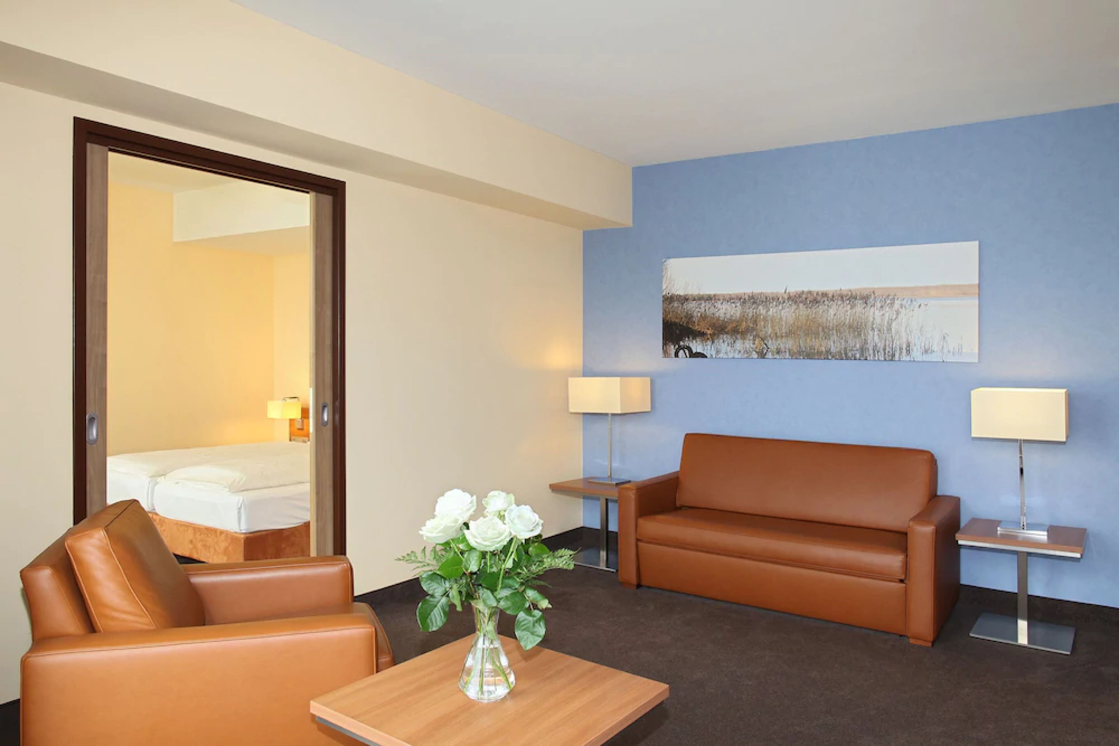 Et hotelværelse med en brun lædersofa, et træbord med en vase med hvide blomster, et billede på væggen og to sengeborde med lamper.