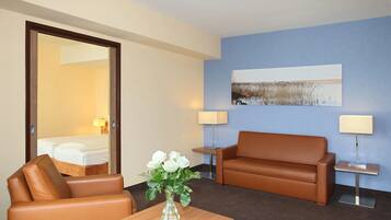 Habitación de hotel con un sofá de cuero marrón, una mesa de centro de madera con un jarrón de flores blancas, un cuadro en la pared y dos lámparas de noche.