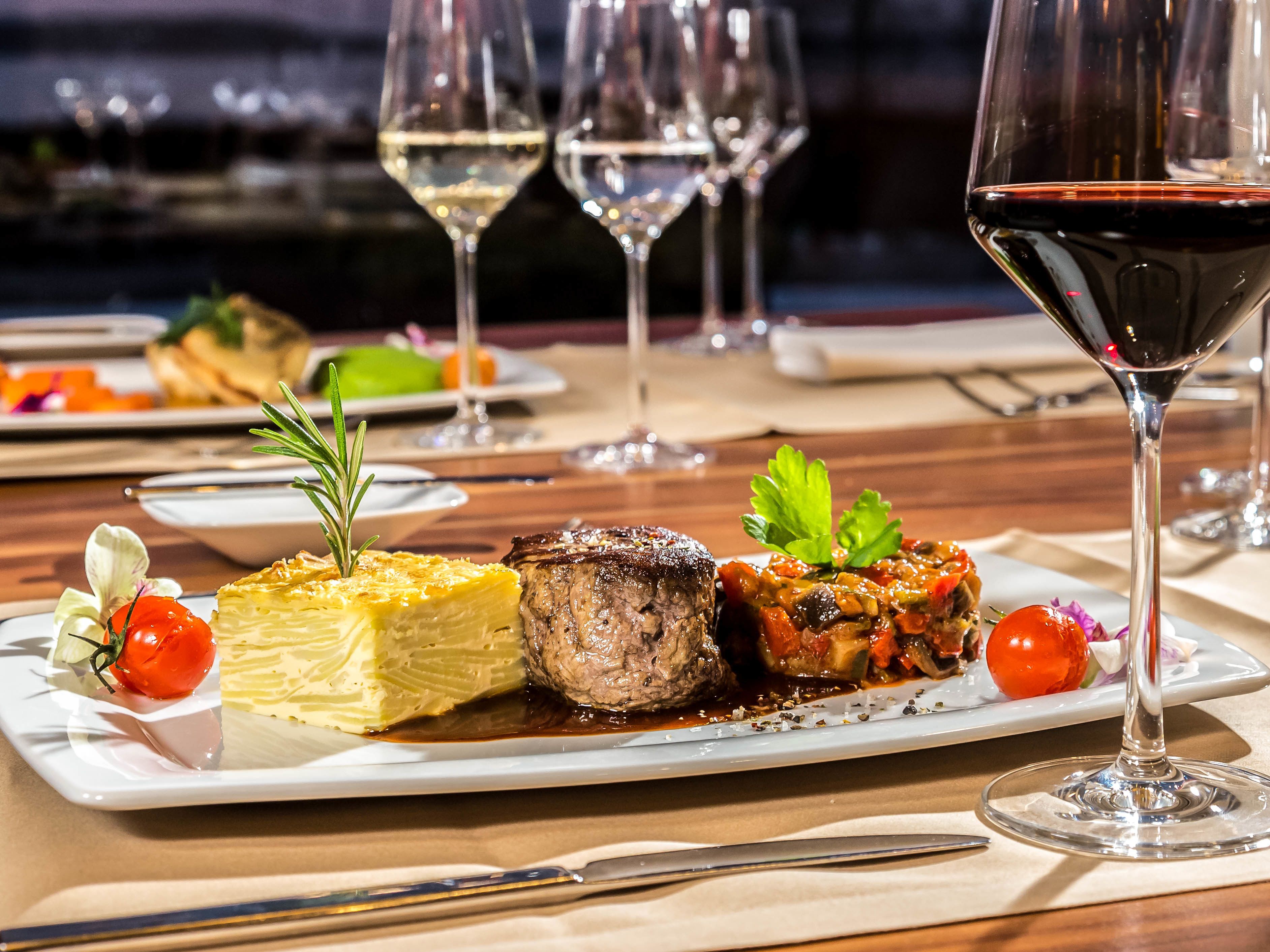 Un tavolo imbandito con diversi piatti, tra cui una bistecca, una terrina di verdure e una quiche, insieme a bicchieri di vino.
