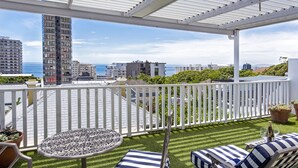 Uma varanda com vista para o oceano, um cercado branco, uma mesa redonda e duas cadeiras de descanso com almofadas listradas.