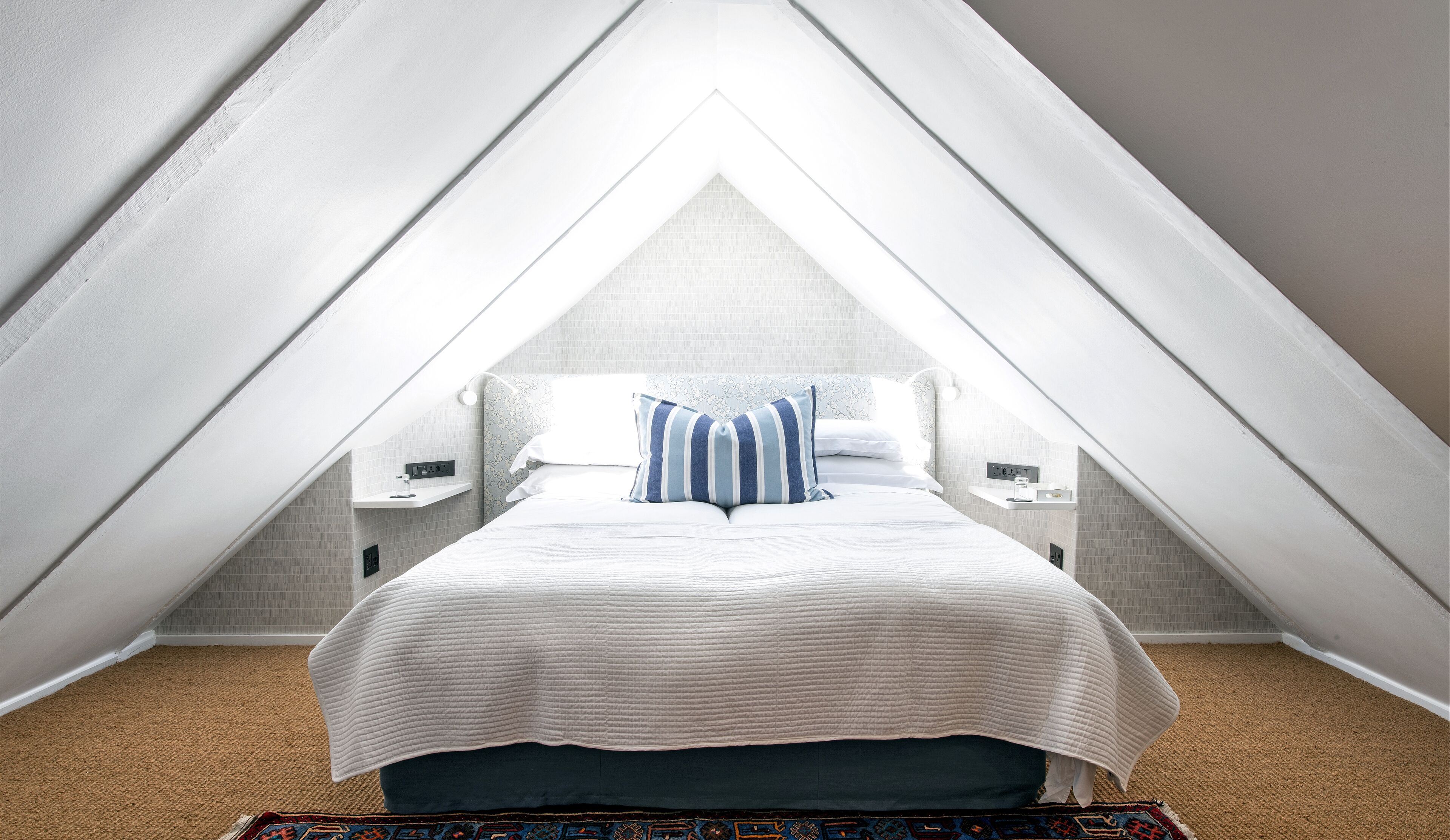 A bedroom with a sloped ceiling, a bed with a white quilt, a blue and white striped pillow, and a nightstand with a lamp.