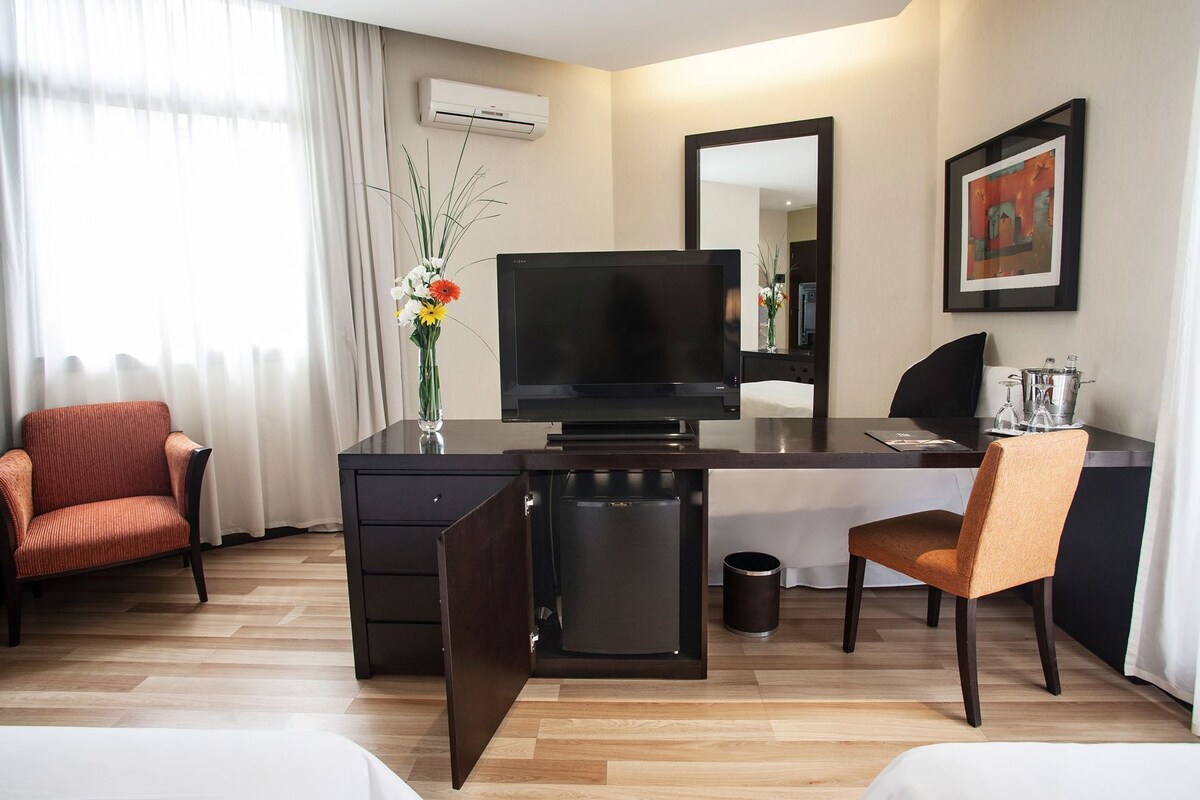 Habitación de hotel con televisión de pantalla plana, escritorio, dos sillas, refrigerador pequeño, cesto de basura y un florero con flores.