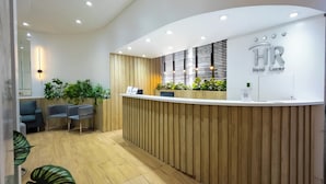 Zona de recepción de hotel con mostrador de madera, área de asientos con sillas y un letrero en la pared.