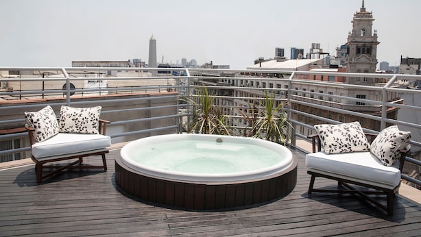 Terraza en la azotea con dos sofás blancos acolchados y un jacuzzi, con vistas a un paisaje urbano con edificios y una torre del reloj.