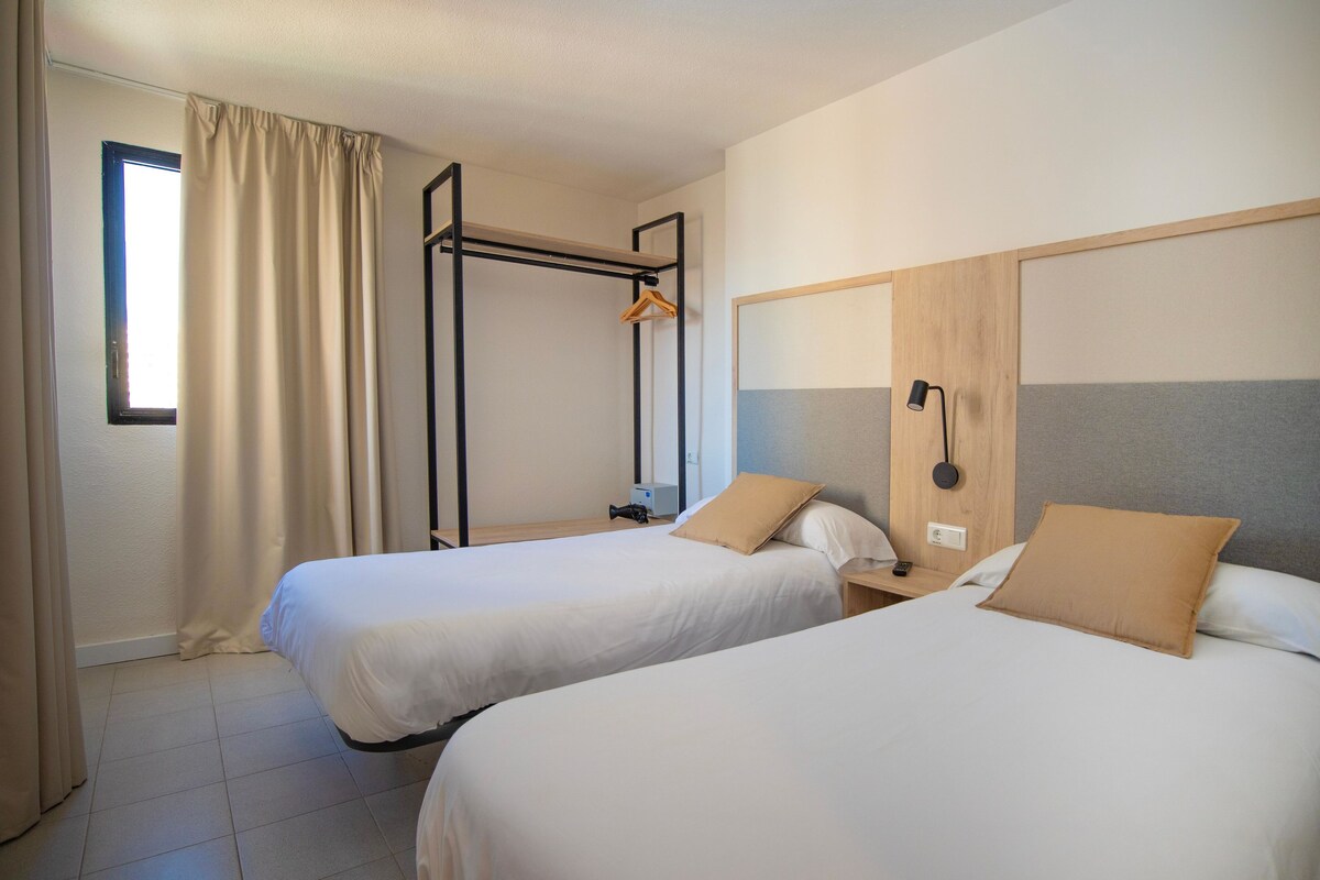 A hotel room with two beds, a wooden headboard, a hanging rack, and a window with curtains.