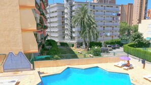 A swimming pool with lounge chairs and a palm tree, surrounded by a multi-story building with balconies. - Apartamentos Dynastic Rooms (Benidorm)