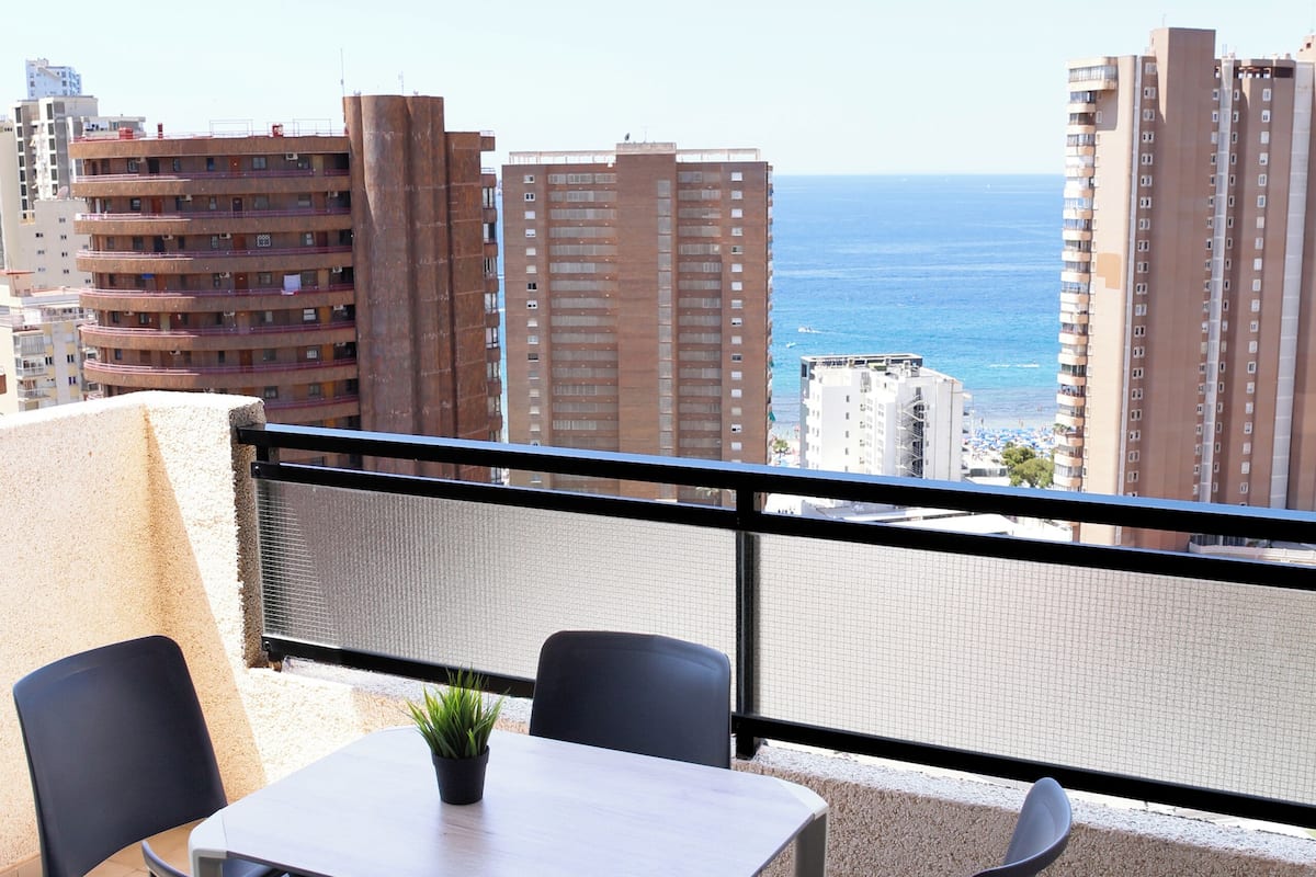 A balcony with a table and chairs, overlooking a cityscape and the sea.