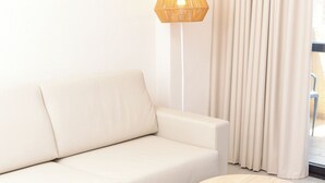 A modern living room with a beige sofa, two round wooden tables, a floor lamp, and a view through a window with curtains. - Apartamentos Dynastic Rooms (Benidorm)