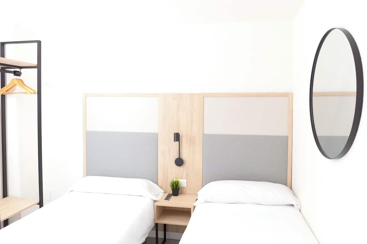 A hotel room with two single beds, a wooden headboard, a bedside table with a plant, a hanging light, and a mirror.