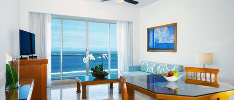 A modern hotel room with a large window offering a view of the ocean, a dining area with a glass table and chairs, a television, and a ceiling fan.