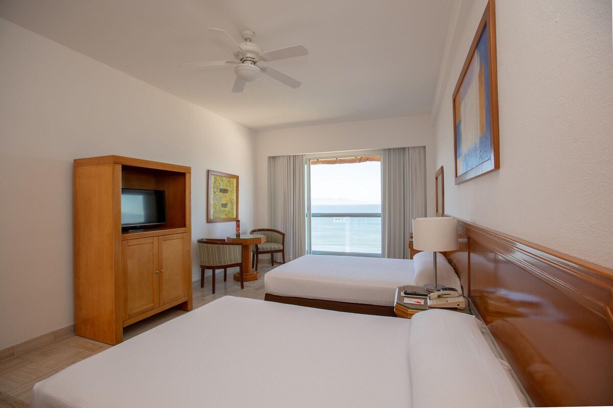 A hotel room with two beds, a wooden cabinet with a television, a desk with a chair, a ceiling fan, and a large window with a view of the sea.