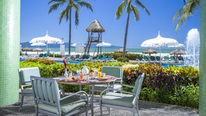 Un espace repas offrant une vue sur la piscine et la plage, entouré de palmiers et de parasols.