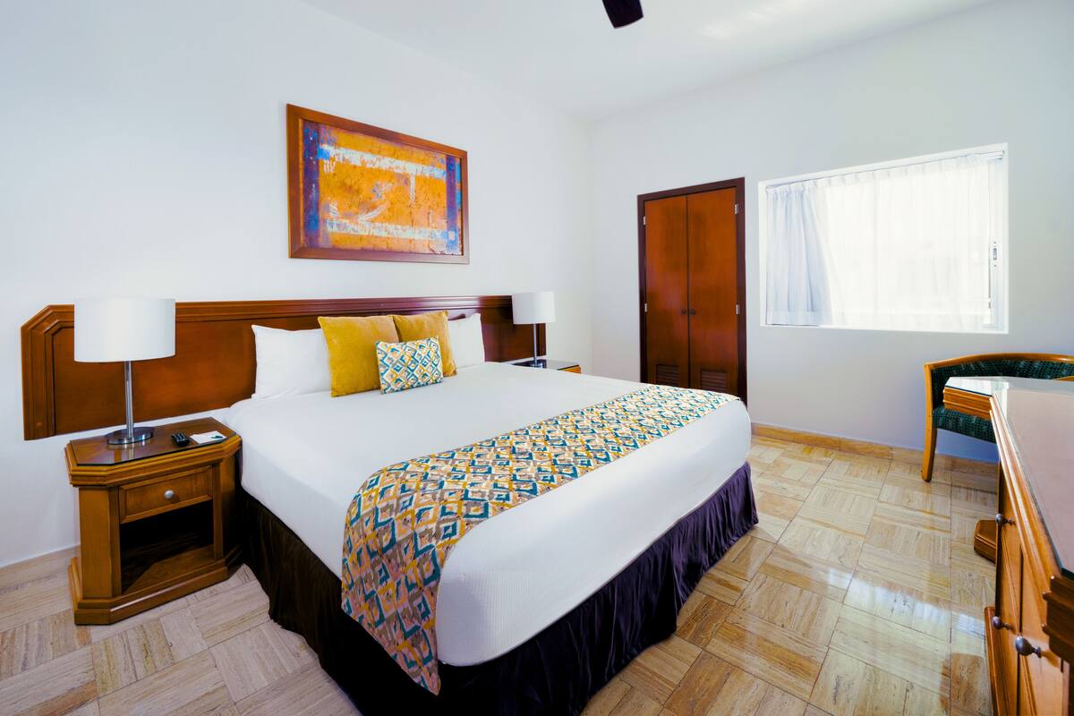 Une chambre d’hôtel avec un grand lit, une tête de lit en bois, une table de chevet avec une lampe, une chaise, un bureau et une fenêtre avec des rideaux.