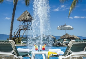 Uma área de piscina com espreguiçadeiras, uma estrutura com telhado de palha e vista desobstruída para o oceano.