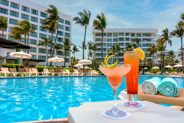 Vista da piscina com dois cocktails, cadeiras de descanso e palmeiras.