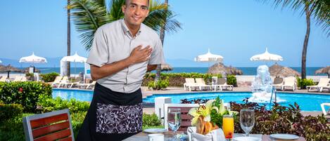 Mesa de jantar posta com pratos, talheres e uma tigela de frutas, com vista para uma piscina e espreguiçadeiras com guarda-sóis.