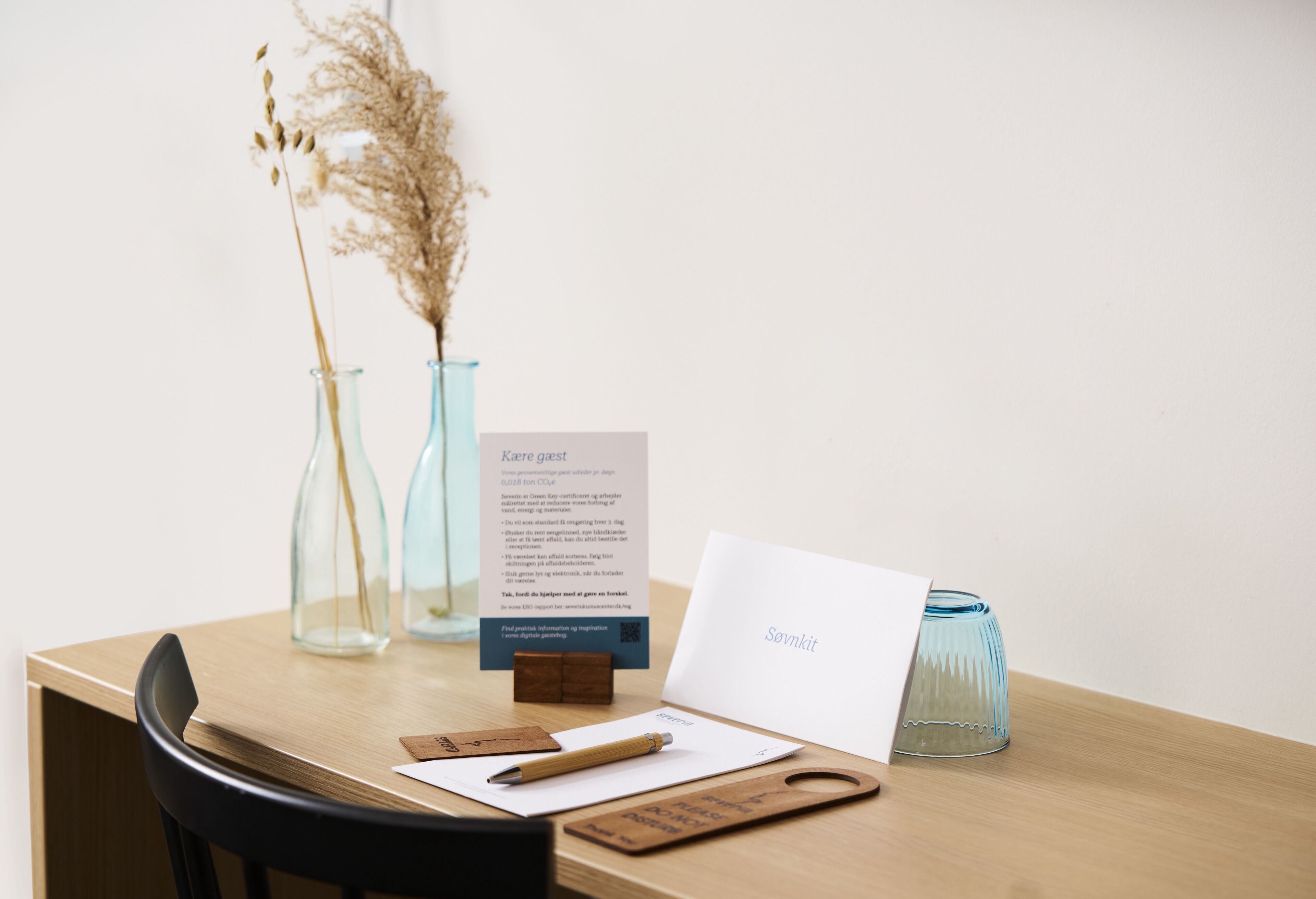 A wooden table with a whiteboard, a wooden cutting board, a pencil, two glass vases with dried plants, and a card with text.