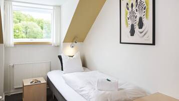 A bedroom with a bed, a bedside table, a nightstand, a window with curtains, a radiator, and a framed zebra artwork on the wall.